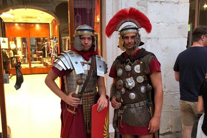 Two individuals in Roman military costumes standing in front of a building with a glass door. 5