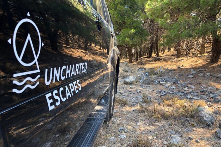 Een voertuig met de tekst 'UNCHARTED ESCAPES' erop, geparkeerd in een bebost gebied met een rotsachtig pad. 1