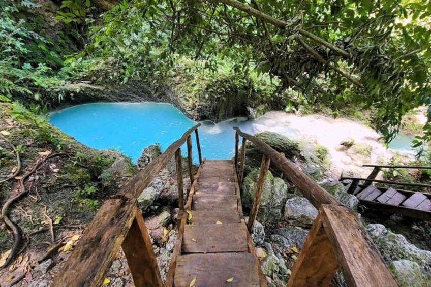 A wooden bridge leading to a turquoise pool surrounded by lush greenery. 1