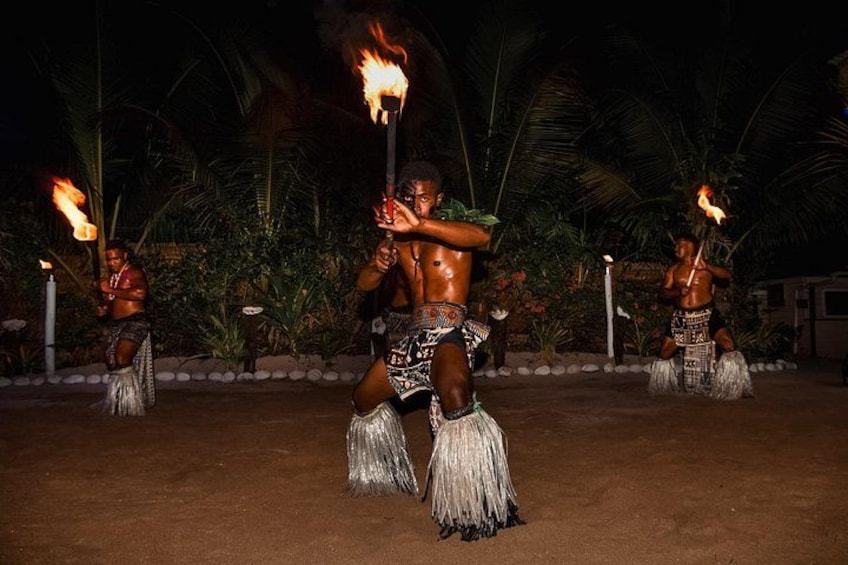 Uno spettacolo culturale notturno con artisti che brandiscono torce fiammeggianti, vestiti con costumi tradizionali, in un ambiente esterno con palme e terreno sabbioso. 3