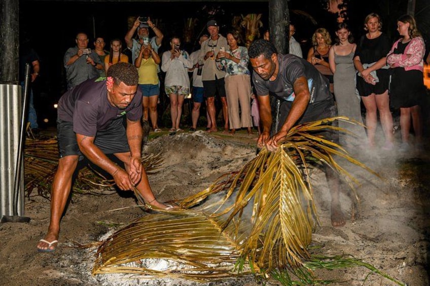 Due uomini preparano un pesce grande su una superficie sabbiosa, mentre una folla osserva. 4