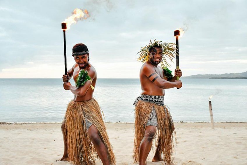 Two people in traditional grass skirts and headdresses, holding flaming tiki torches on a beach with a body of water and distant land in the background. 2