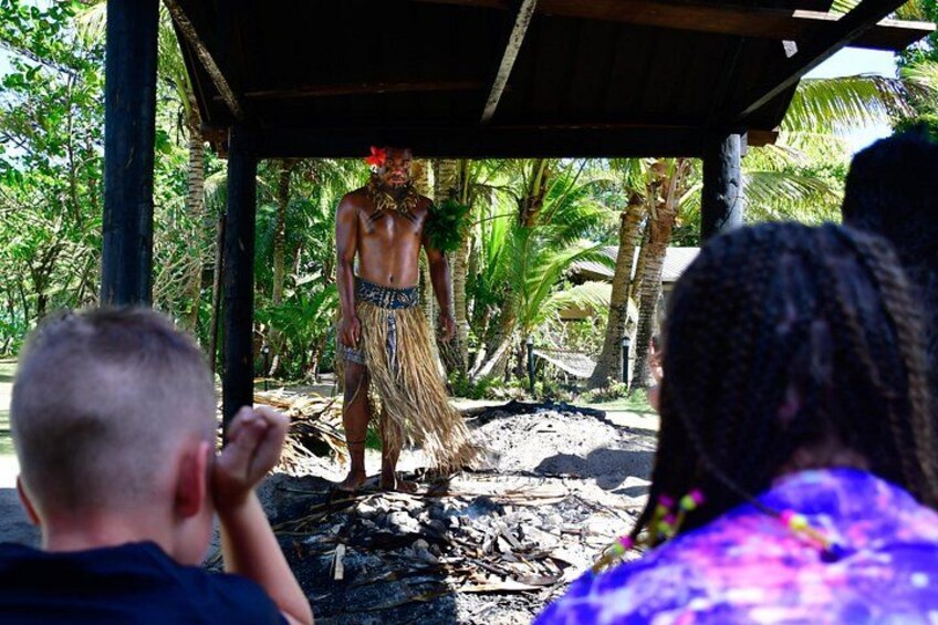 Une personne vêtue d’un costume traditionnel, debout sous un abri, avec un paysage tropical en arrière-plan. 2