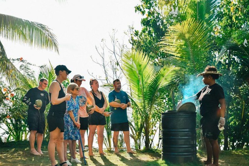 A group of people outdoors near a large black barrel with smoke, surrounded by tropical vegetation. 4