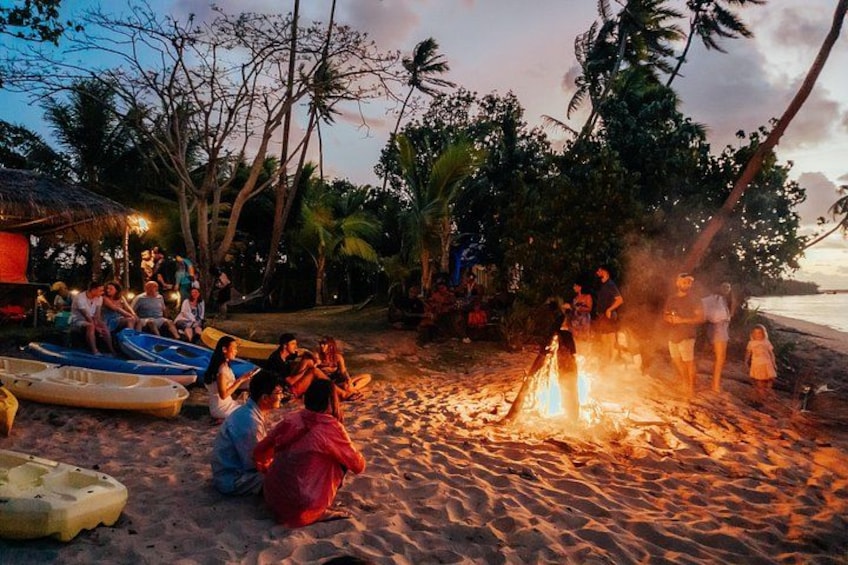 A beach scene with people gathered around a fire, boats, and palm trees. 2