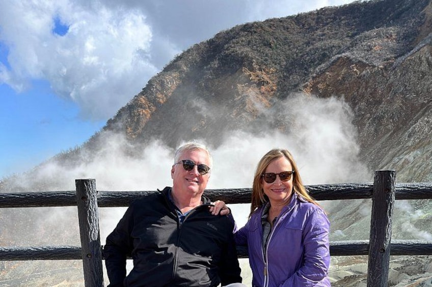 Two people standing by a railing with a mountainous landscape and steam in the background. 1