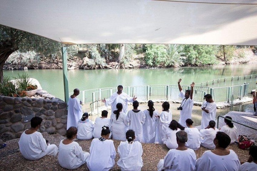 Un groupe de personnes vêtues de longues robes blanches est assis et se tient debout à l’extérieur, près d’une étendue d’eau. On aperçoit en arrière-plan un mur de pierre et de la végétation. 1