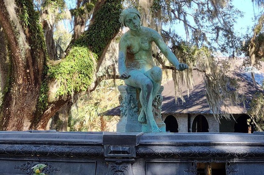 A bronze statue of a woman seated on a rock, surrounded by trees and a building with arched windows. 5