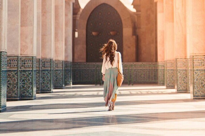 A person walking in a sunlit architectural corridor with ornate metal railings and a large arched doorway. 3