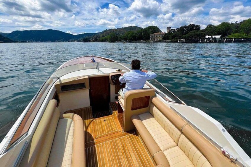 Een boot met een stuurman, houten dek en een uitzicht over een meer met heuvels en gebouwen op de achtergrond. 2
