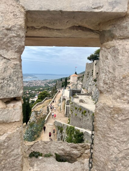 Un chemin de pierre mène à un bâtiment historique, orné d’un drapeau, qui surplombe un paysage urbain côtier. 2