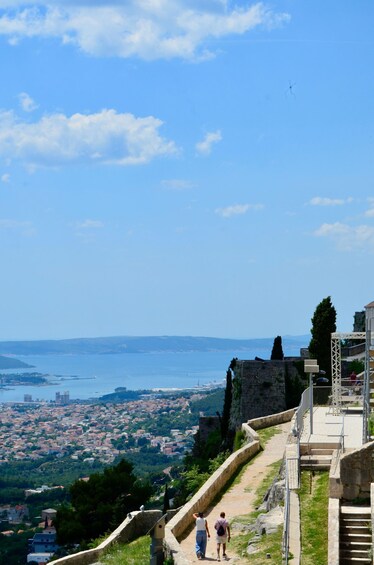 A coastal cityscape with a clear view of the sea, a historic stone wall, and a paved walkway. 1