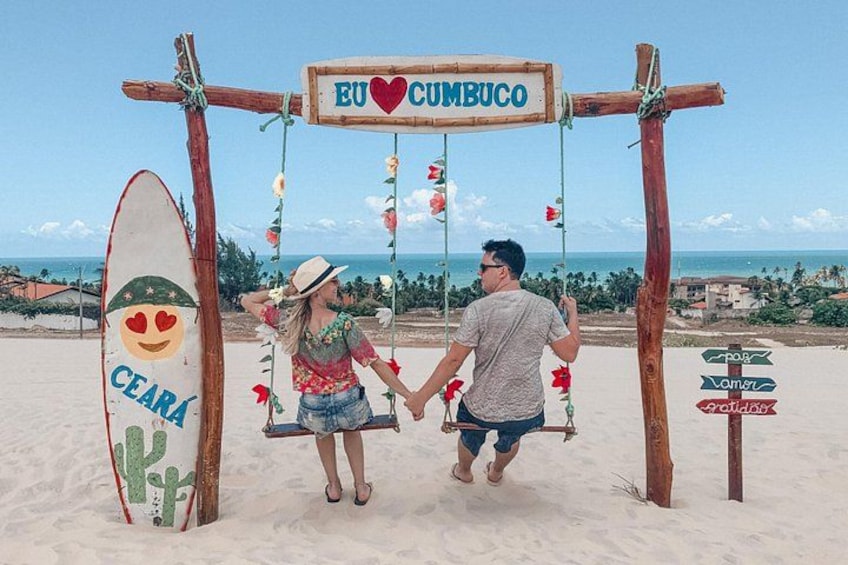 Un columpio de madera con un letrero que dice 'EU CUMBUCO' y una tabla de surf con un diseño de cactus. 1
