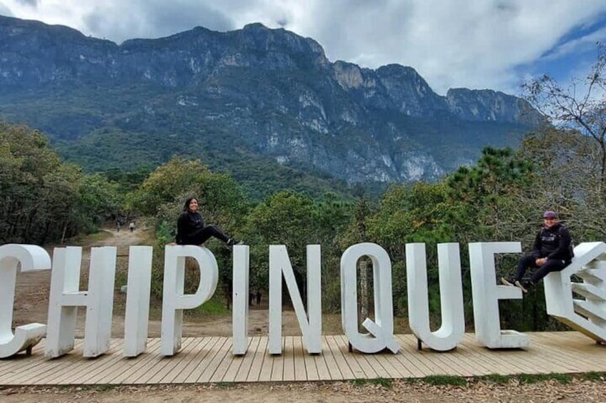 Two people sitting on large white letters spelling "CHIPINQUE" with a mountainous landscape in the background. 1