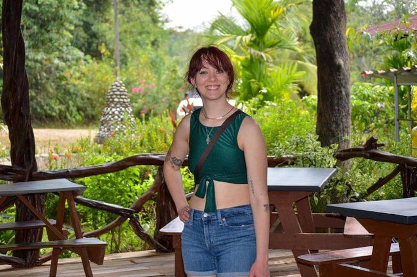 A person standing outdoors with a green top and blue jeans, surrounded by wooden tables and chairs, with greenery and a tree in the background. 4