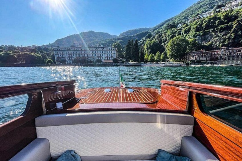 A boat with a wooden deck, a cushioned seating area, and a view of a lake with buildings and hills in the background. 1