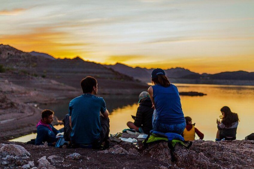 A group of people sitting by a lake at sunset with mountains in the background. 4