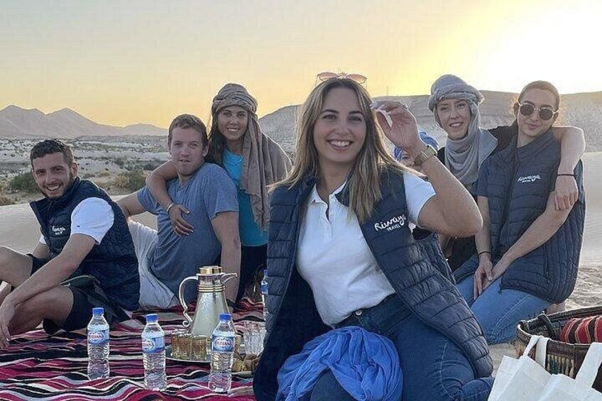 Un groupe de personnes est assis sur une couverture à l’extérieur, avec un paysage désertique en arrière-plan. 1