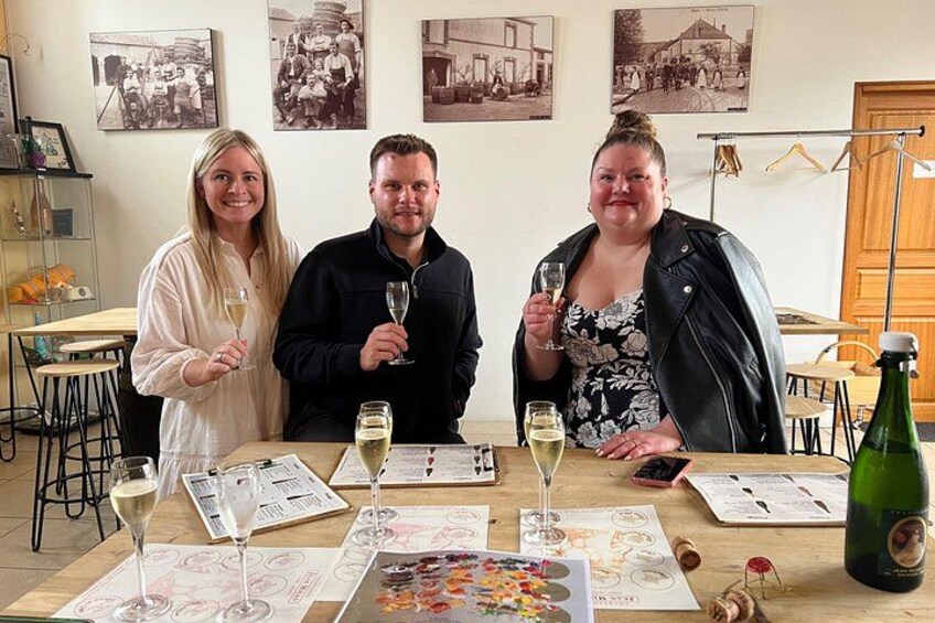 Tres personas sentadas a una mesa con copas de vino, menús y una botella de vino. 3