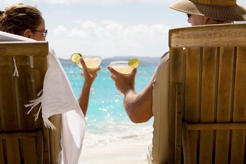 Dos personas sentadas en sillas de playa de madera mirando al océano, sosteniendo bebidas con gajos de limón. 2
