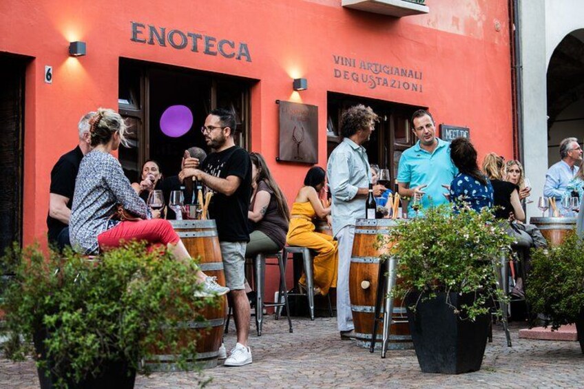 Personas sentadas y de pie frente a un edificio con barriles, un letrero que dice 'ENOTECA' y un cartel anunciando 'VINI ARUGIANALI DEGUSTAZIONI'. 1