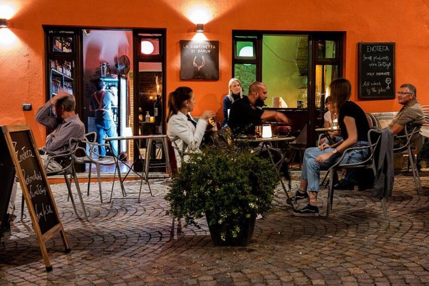 Un café al aire libre con gente sentada en mesas, una planta en maceta y un cartel. 4