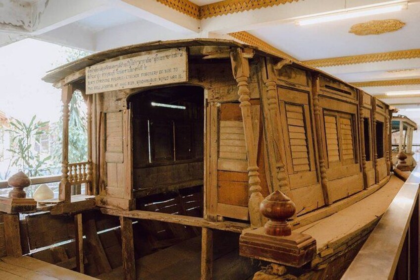 A wooden ship model with a sign, displayed indoors with a railing and decorative elements. 5