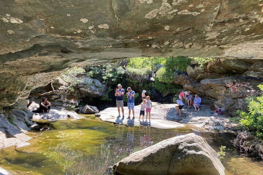 A group of people standing and sitting around a small stream in a rocky area with greenery. 2