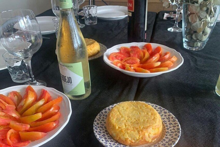 Una mesa con una botella de vino blanco, un plato de tomates en rodajas y un pastelito redondo. 1