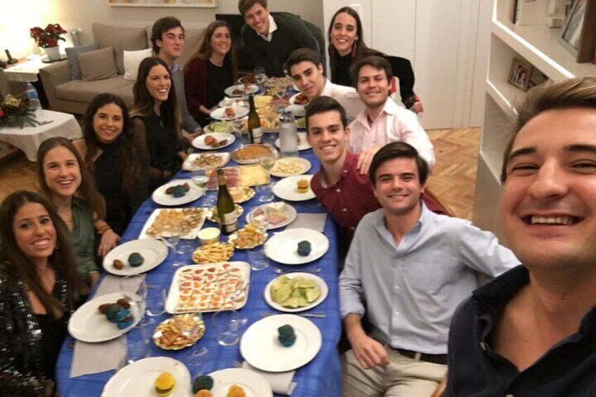Un grupo de personas sentadas alrededor de una mesa con varios platos, en una habitación que tiene un sofá y una estantería. 3