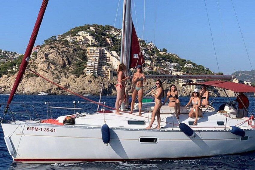 A sailboat with people on deck, a rocky coastline with buildings, and clear blue water. 5