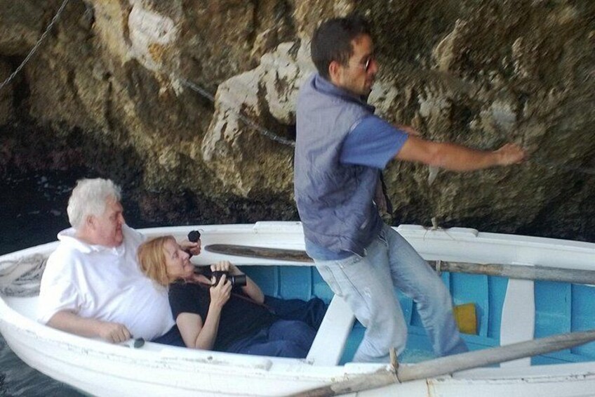Un petit bateau avec trois personnes à bord : une personne est debout et deux sont assises, sur une plage rocheuse. 1