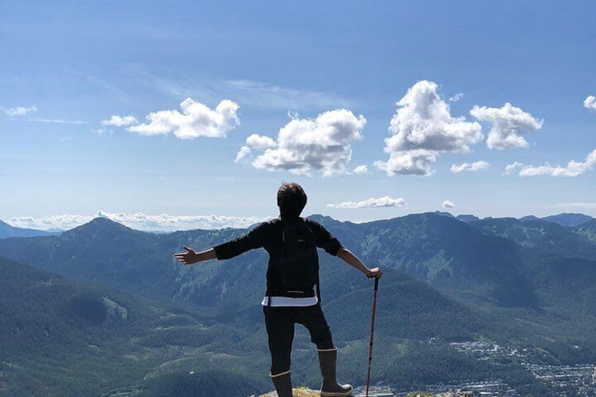 A person standing on a mountain peak with arms outstretched, wearing a dark jacket, shorts, and boots, and holding a walking stick. 1