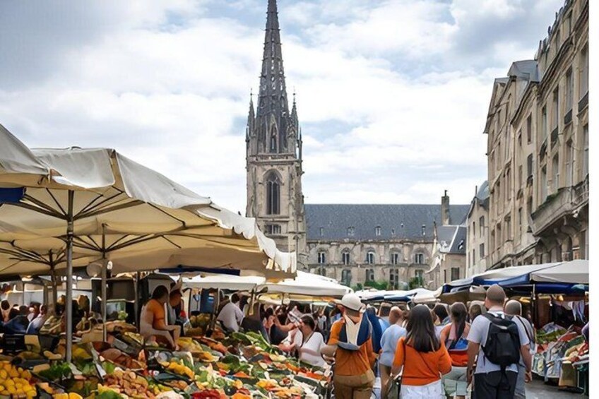 A bustling outdoor market with people shopping, surrounded by historic buildings and a prominent church spire. 1
