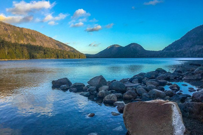 Un lac paisible, avec des rochers au premier plan, entouré de collines boisées sous un ciel dégagé. 2