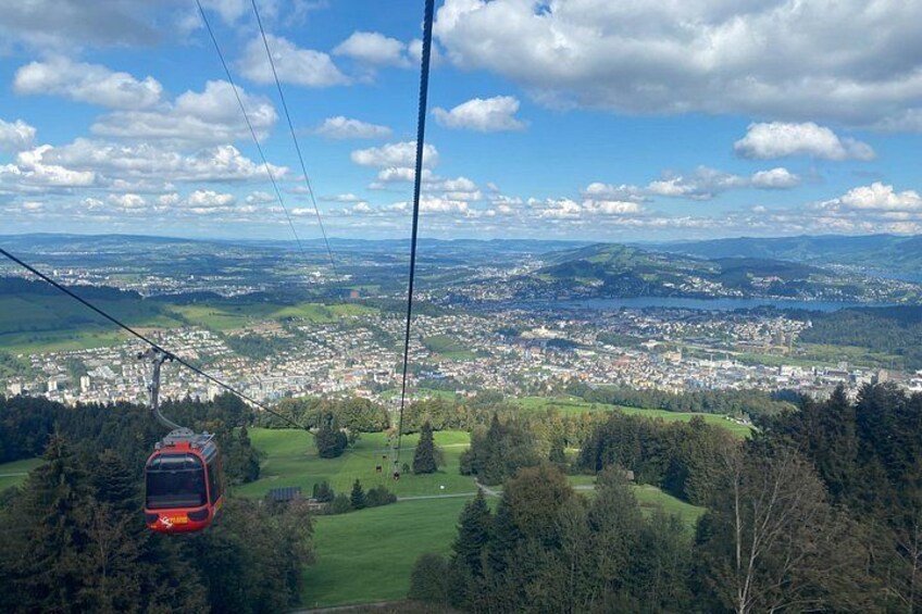 Una funivia che si muove sopra un paesaggio con una città in basso, circondata da vegetazione e colline. 3