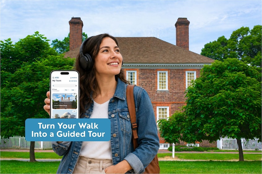 A person holding a smartphone with a travel app interface, standing in front of a brick building with a white fence and greenery. 3
