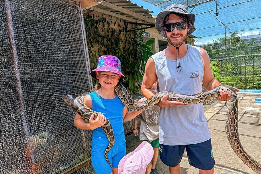 Una persona sosteniendo una serpiente grande, otra persona con sombrero, y una cerca al fondo. 4