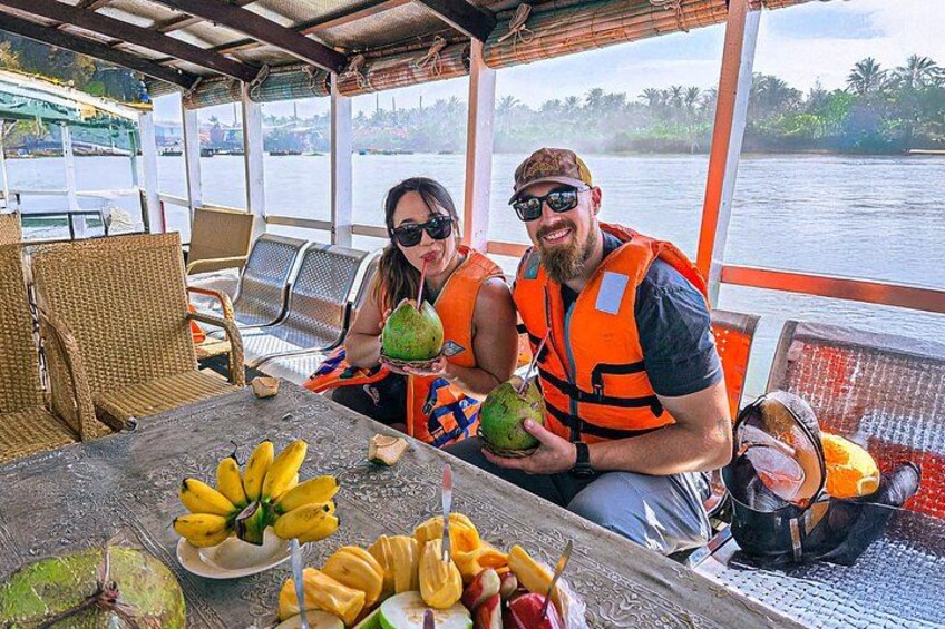 Two people on a boat, one holding a coconut and the other holding a drink. There are coconuts and bananas on the table. 3