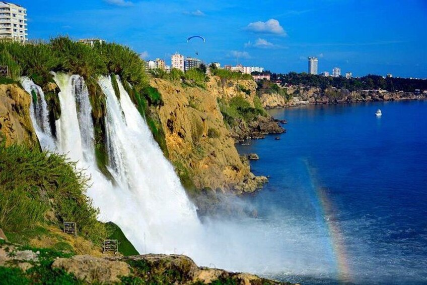 Een waterval aan de kust met een regenboog, gebouwen op de achtergrond en een heldere lucht. 3