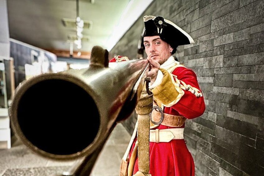 A person in a red uniform with a black hat holding a large brass cannon. 2