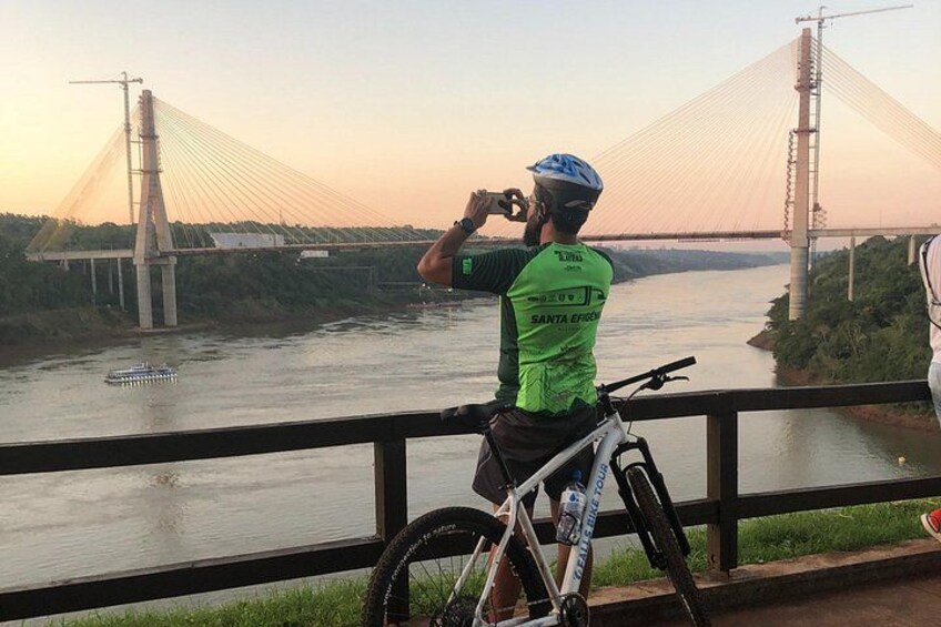 A person in a green cycling jersey taking a photo of a bridge. 3
