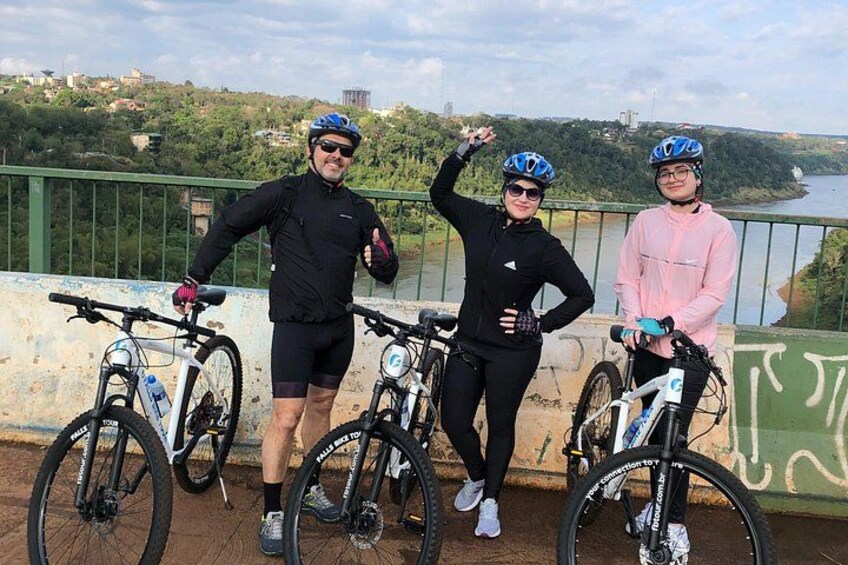 Three cyclists on a bridge with bicycles, a river, and a hilly landscape in the background. 2