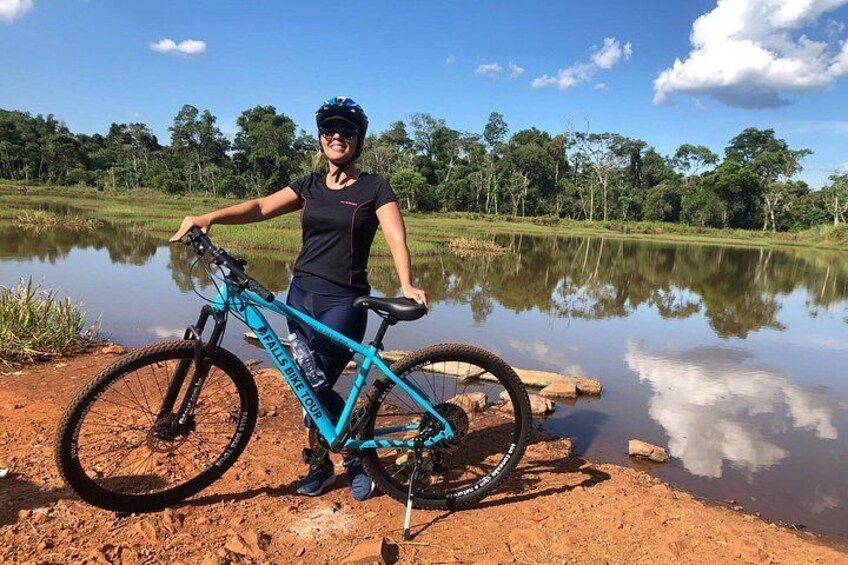 A person standing next to a mountain bike with a water body and trees in the background. 1