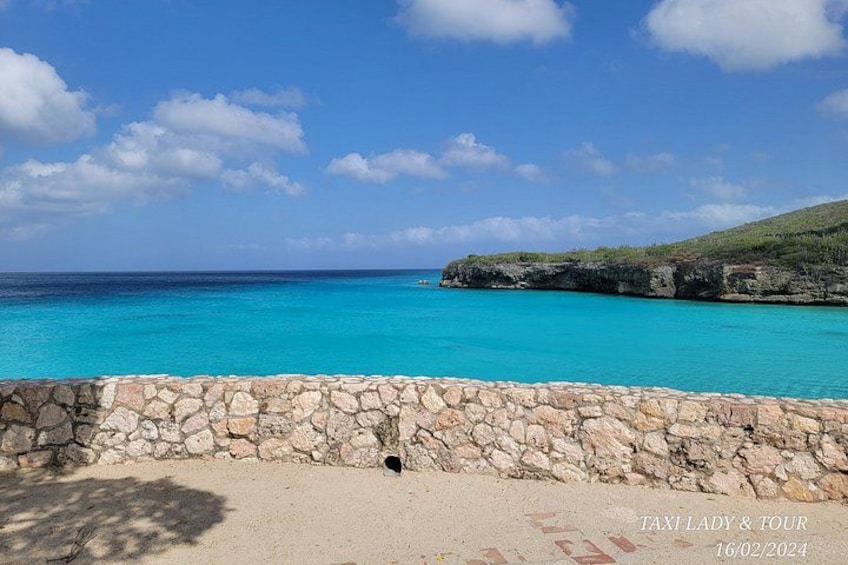 Un paysage côtier avec un mur de pierre, une eau turquoise et limpide, et un affleurement rocheux lointain sous un ciel bleu. 1