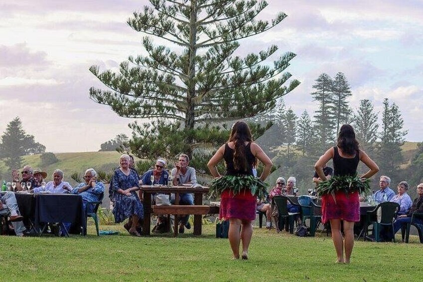 Two women in traditional attire walking in a grassy field with a group of seated people watching. 4