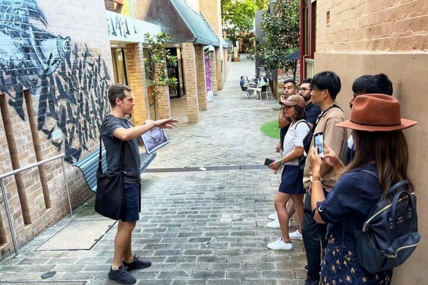 A person pointing at a wall with graffiti, standing on a cobblestone street with a group of people observing. 1