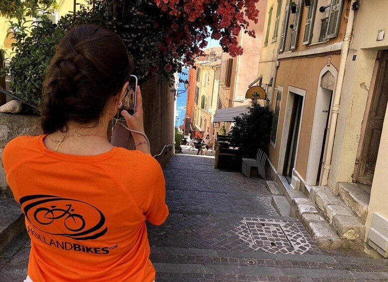 A person taking a photo on a cobblestone street with buildings and a tree with pink flowers. 3