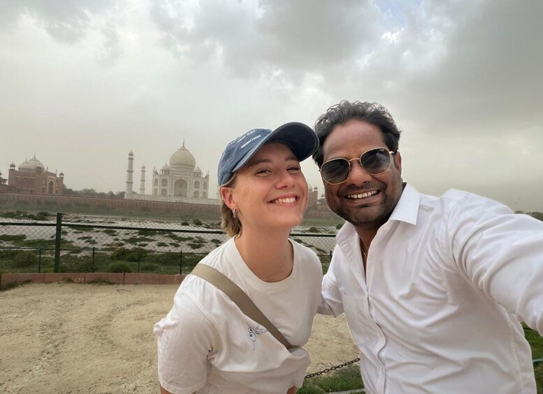 A person in a white shirt and another in a white top with a cap, standing in front of the Taj Mahal. 1