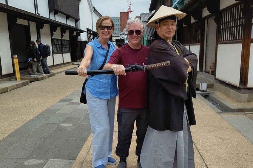A street with traditional architecture, a person in a straw hat, and another holding a sword. 2